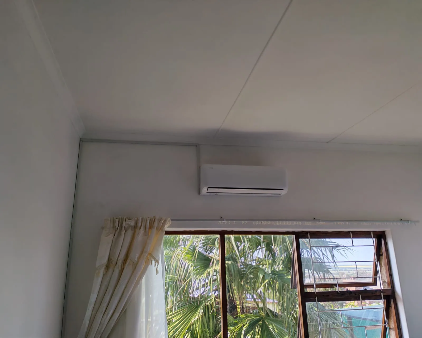 Indoor split air-conditioning unit installed near a window and curtain rail.