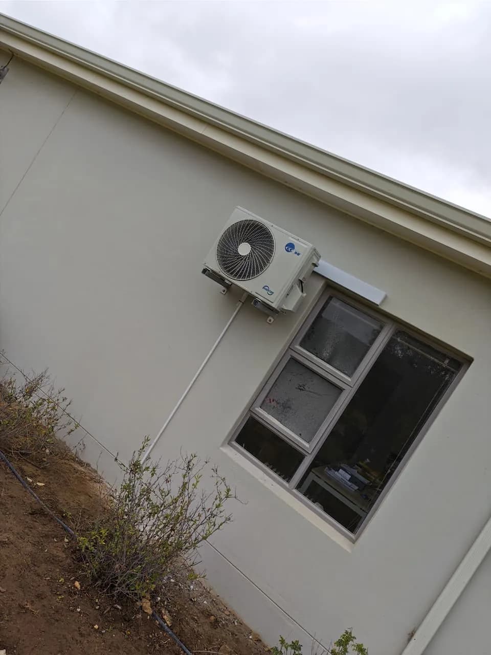 Angled exterior view of an air-conditioning condenser and enclosed pipework beside a window.